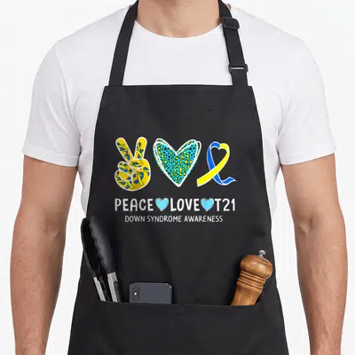Cure ribbon apron - Peace hand, leopard heart, and ribbon with PEACE LOVE T21 text in yellow blue and white — shown on a black cotton canvas apron against a white background
