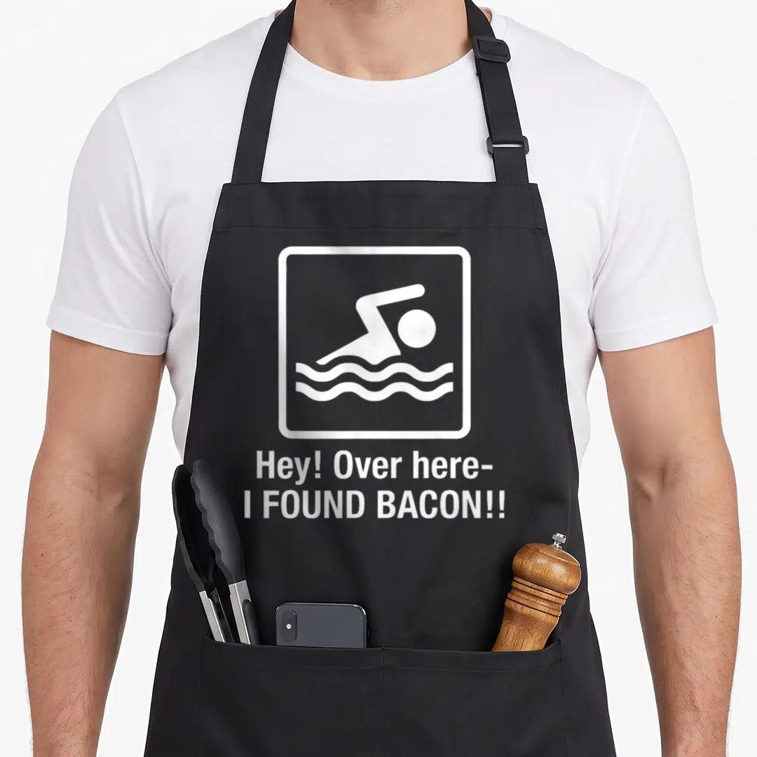 Found bacon apron - White swimmer pictogram in square frame above text Hey! Over here- I FOUND BACON!! — shown on a black cotton canvas apron against a white background