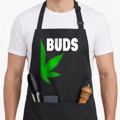 Bud couple apron - Green cannabis leaf illustration beside white BUDS text — shown on a black cotton canvas apron against a white background