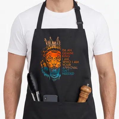 Approval king apron - Crowned orange and blue watercolor lion head with stacked text I'm An Gemini King Your Approval — shown on a black cotton canvas apron against a white background