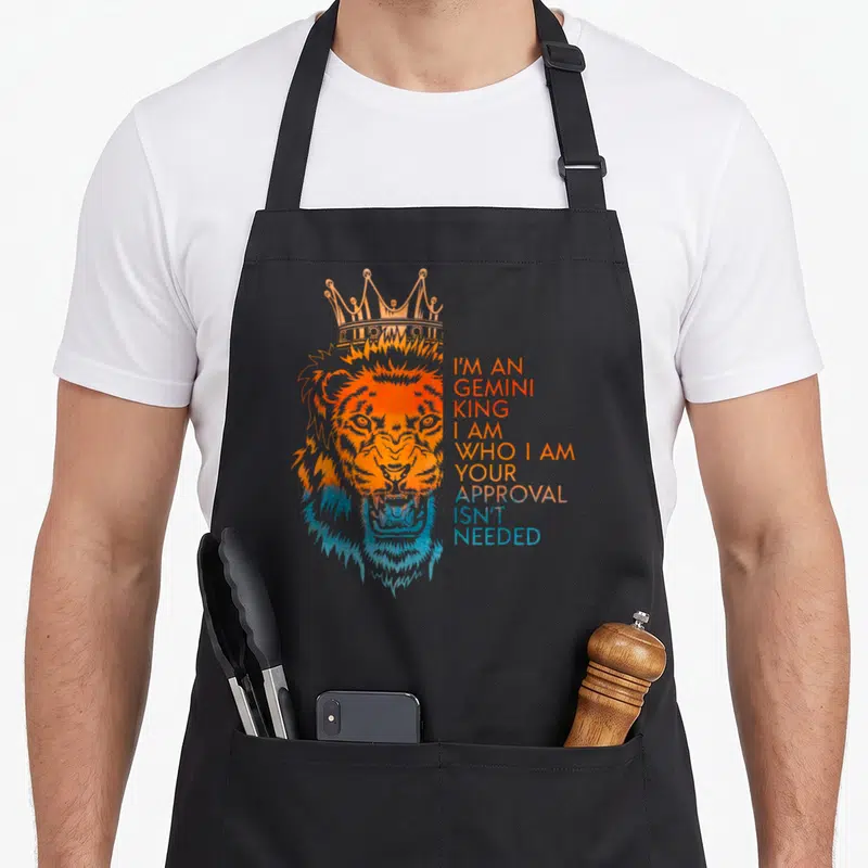 Approval king apron - Crowned orange and blue watercolor lion head with stacked text I'm An Gemini King Your Approval — shown on a black cotton canvas apron against a white background
