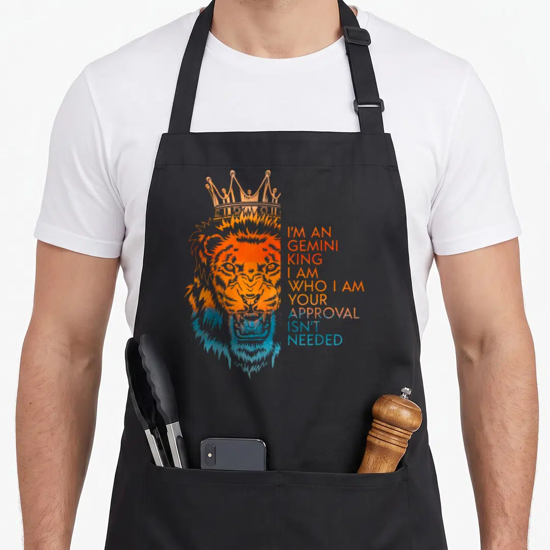 Approval king apron - Crowned orange and blue watercolor lion head with stacked text I'm An Gemini King Your Approval — shown on a black cotton canvas apron against a white background