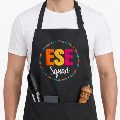 Ese art teacher apron - ESE Squad text surrounded by LOVING FUN COMPASSIONATE words and hearts in orange yellow pink — shown on a black cotton canvas apron against a white background