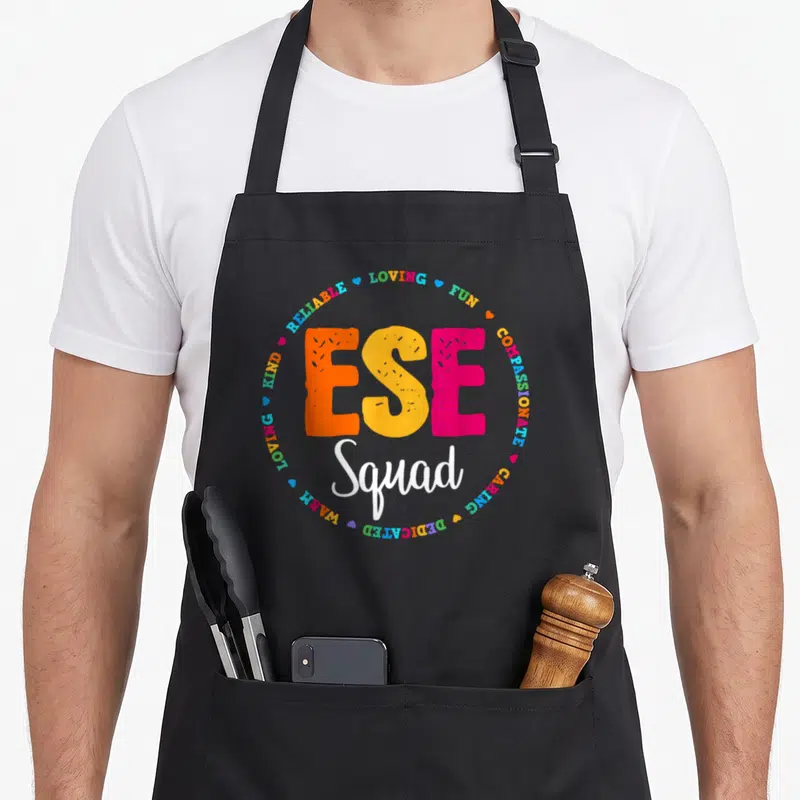 Ese art teacher apron - ESE Squad text surrounded by LOVING FUN COMPASSIONATE words and hearts in orange yellow pink — shown on a black cotton canvas apron against a white background