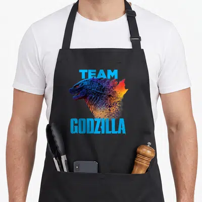 Kong novelty apron - Realistic Godzilla head profile between blue TEAM and GODZILLA text with gradient scales — shown on a black cotton canvas apron against a white background
