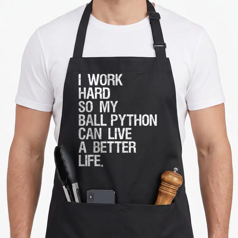 Ball hard apron - White stacked distressed text reading I WORK HARD SO MY BALL PYTHON CAN LIVE A BETTER LIFE — shown on a black cotton canvas apron against a white background