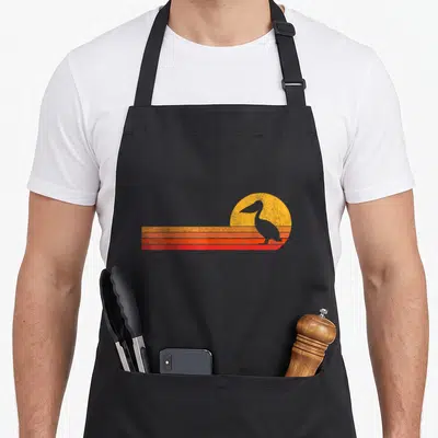 Vintage pelican apron - Retro pelican silhouette against yellow sun with orange and red horizontal stripes — shown on a black cotton canvas apron against a white background