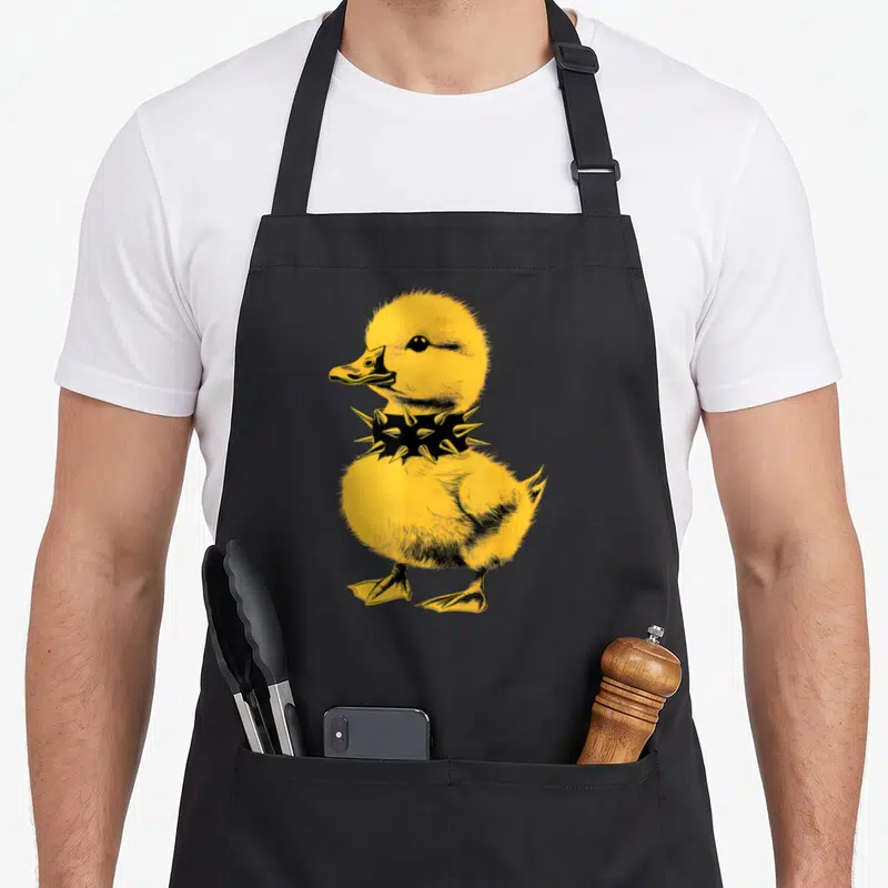 Rocker punk rock apron - Yellow cartoon duckling wearing a black spiked collar — shown on a black cotton canvas apron against a white background