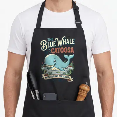 Whale centennial apron - Cartoon blue whale in pond with The Blue Whale of Catoosa and Mother Road Centennial 1926-2026 — shown on a black cotton canvas apron against a white background
