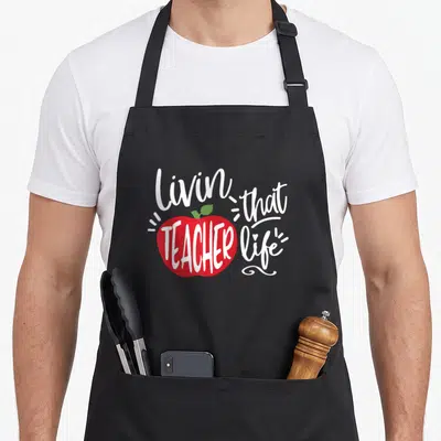 Funny teacher apron - Red apple with white script text Livin that TEACHER life and small green leaf accent — shown on a black cotton canvas apron against a white background