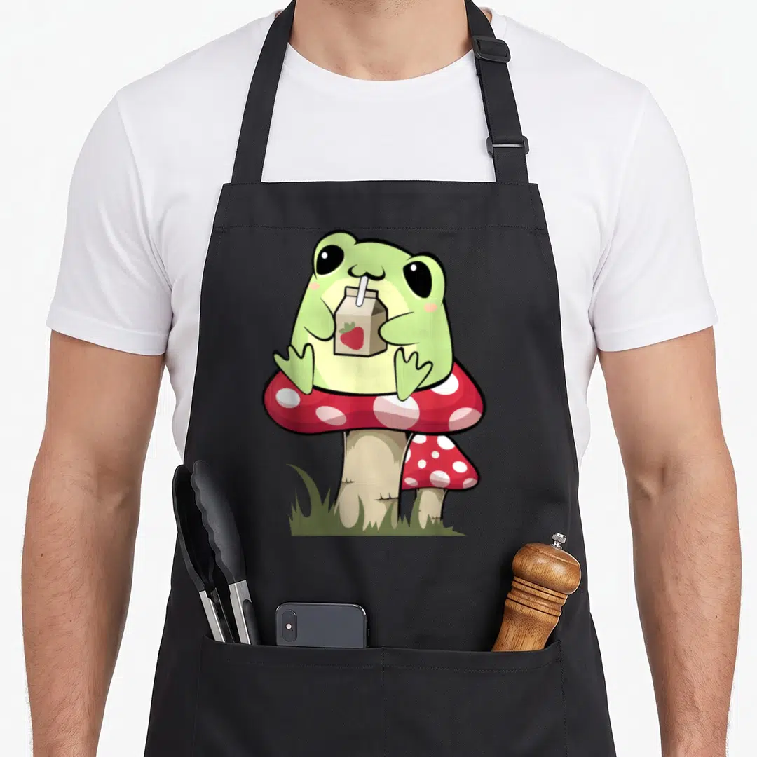 Mycology cottagecore apron - Cartoon green frog drinking strawberry milk on red and white spotted mushroom with grass — shown on a black cotton canvas apron against a white background