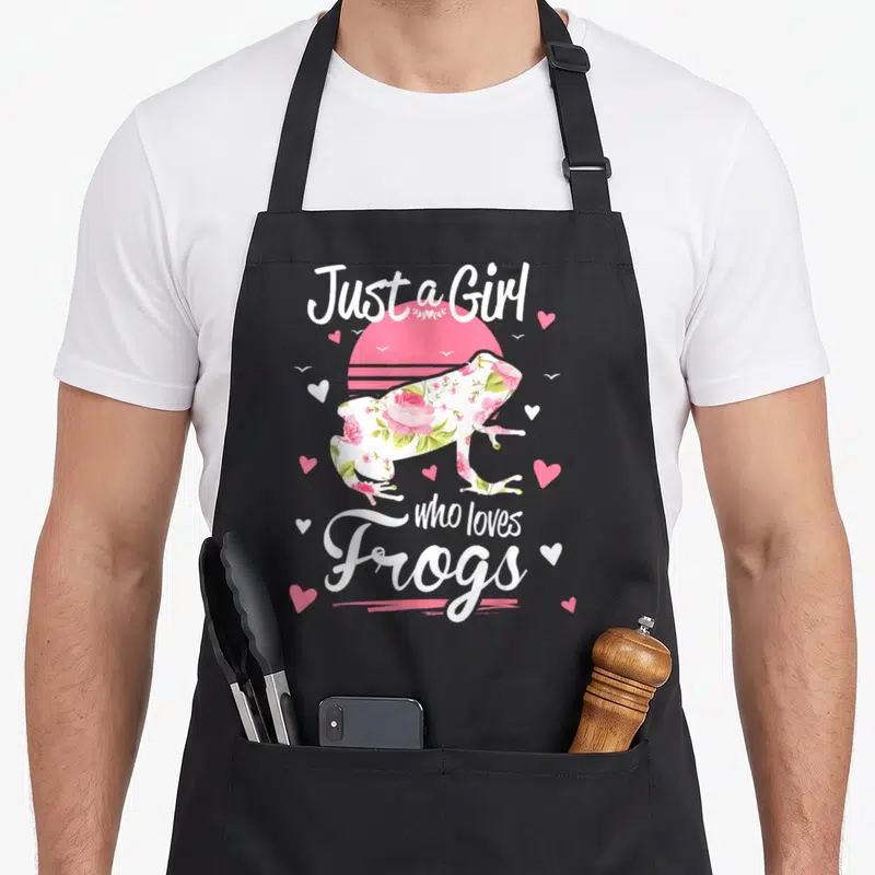 Funny animal frog aprons - Floral frog silhouette over pink sun with white cursive text and scattered hearts — shown on a black cotton canvas apron against a white background