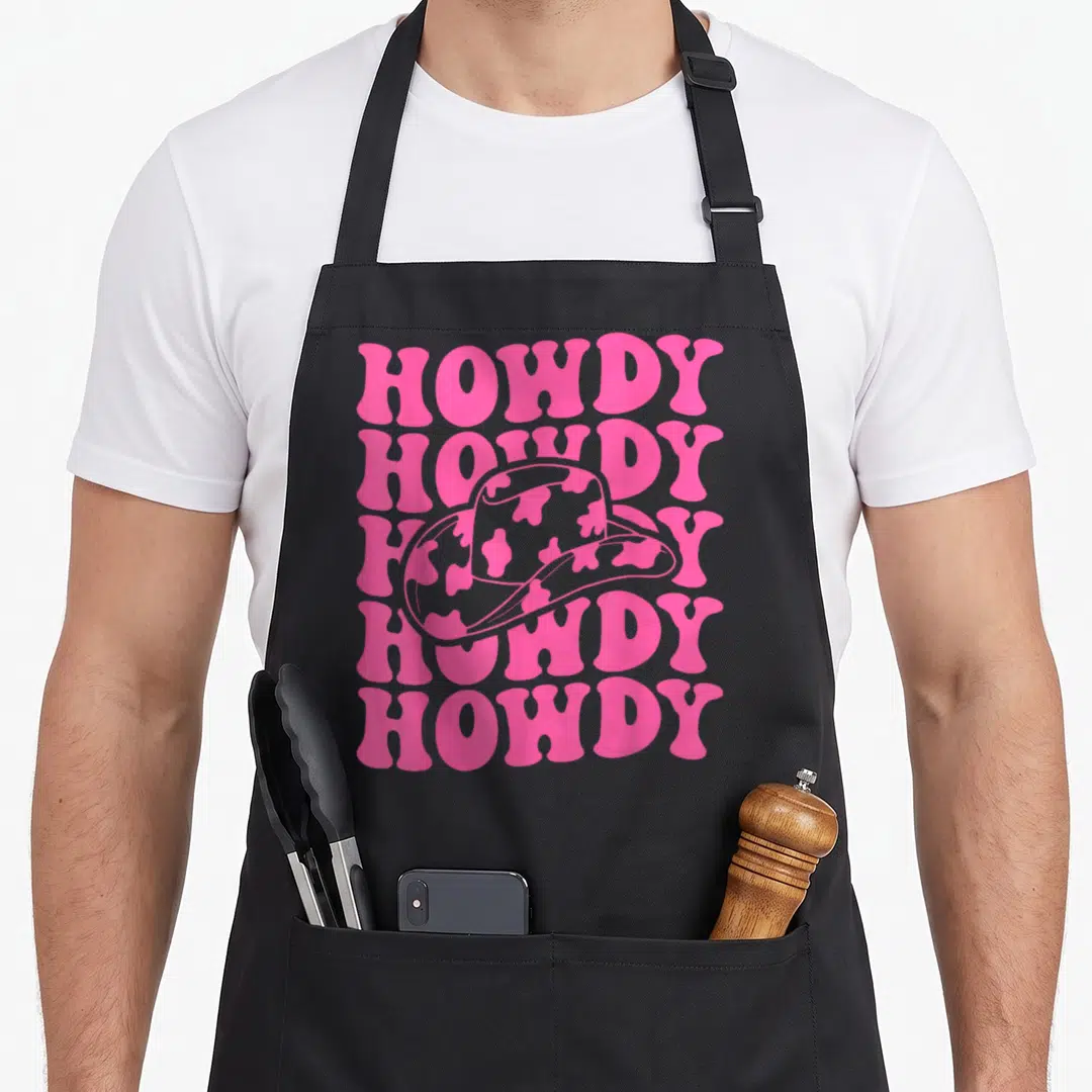 Rodeo southern aprons - Pink stacked HOWDY text with cow-print cowboy hat on grey background — shown on a black cotton canvas apron against a white background