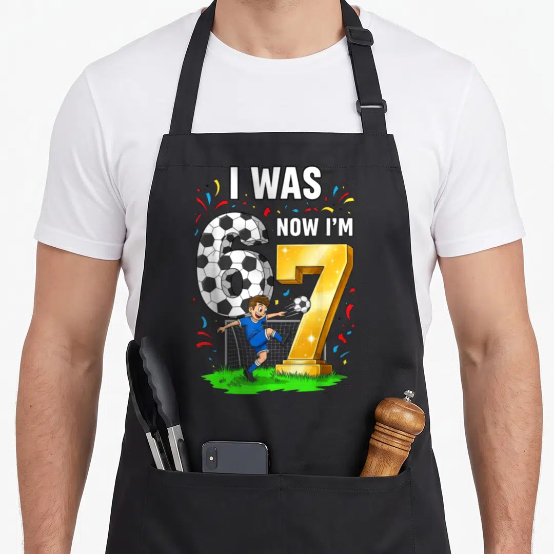 Six seven birthday apron - Cartoon boy kicking soccer ball next to patterned 6 and gold 7 with confetti — shown on a black cotton canvas apron against a white background