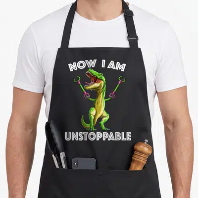 Unstoppable t-rex apron - Green cartoon T-Rex holding pink tool grippers between arched white text NOW I AM UNSTOPPABLE — shown on a black cotton canvas apron against a white background