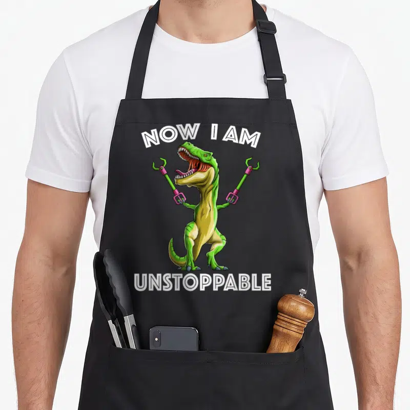 Unstoppable t-rex apron - Green cartoon T-Rex holding pink tool grippers between arched white text NOW I AM UNSTOPPABLE — shown on a black cotton canvas apron against a white background
