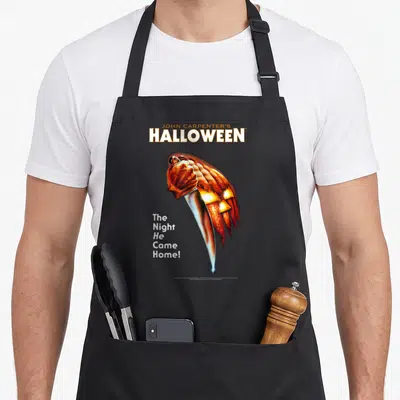 Key halloween apron - Hand holding knife stabbing pumpkin with Halloween title text in orange white silver — shown on a black cotton canvas apron against a white background