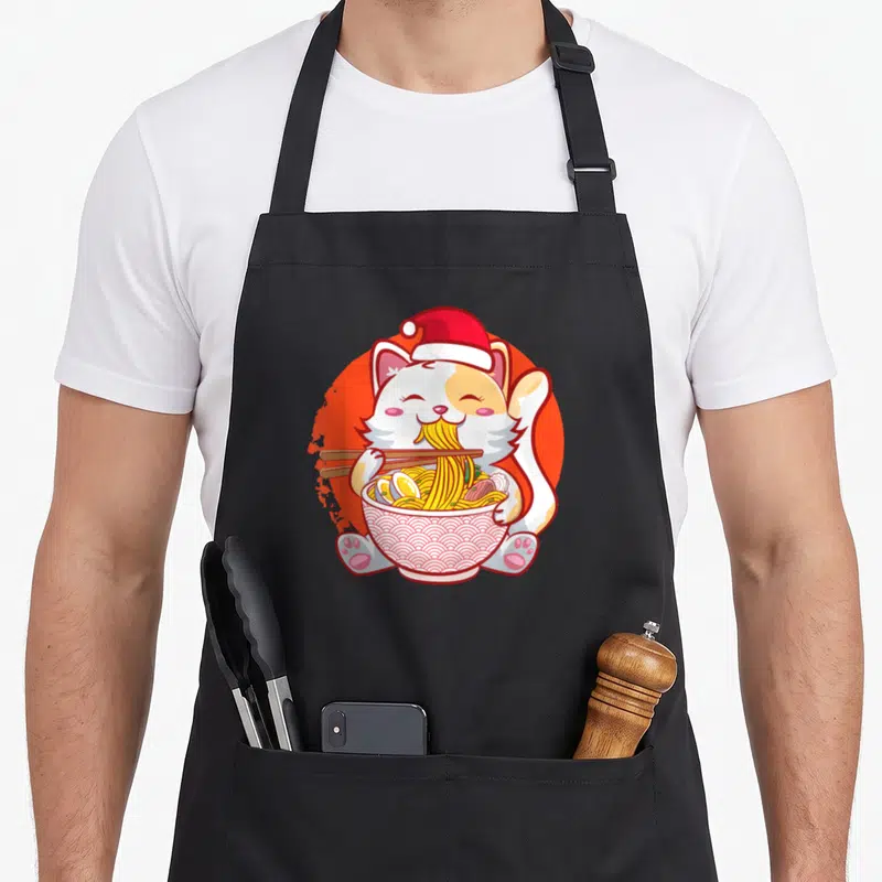 Ramen anime mom apron - Cartoon cat in Santa hat eating ramen noodles with chopsticks on orange circle — shown on a black cotton canvas apron against a white background