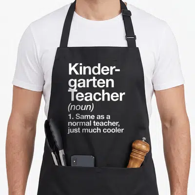 Definition kindergarten teacher apron - White stacked text reading Kinder-garten Teacher noun same as normal teacher — shown on a black cotton canvas apron against a white background