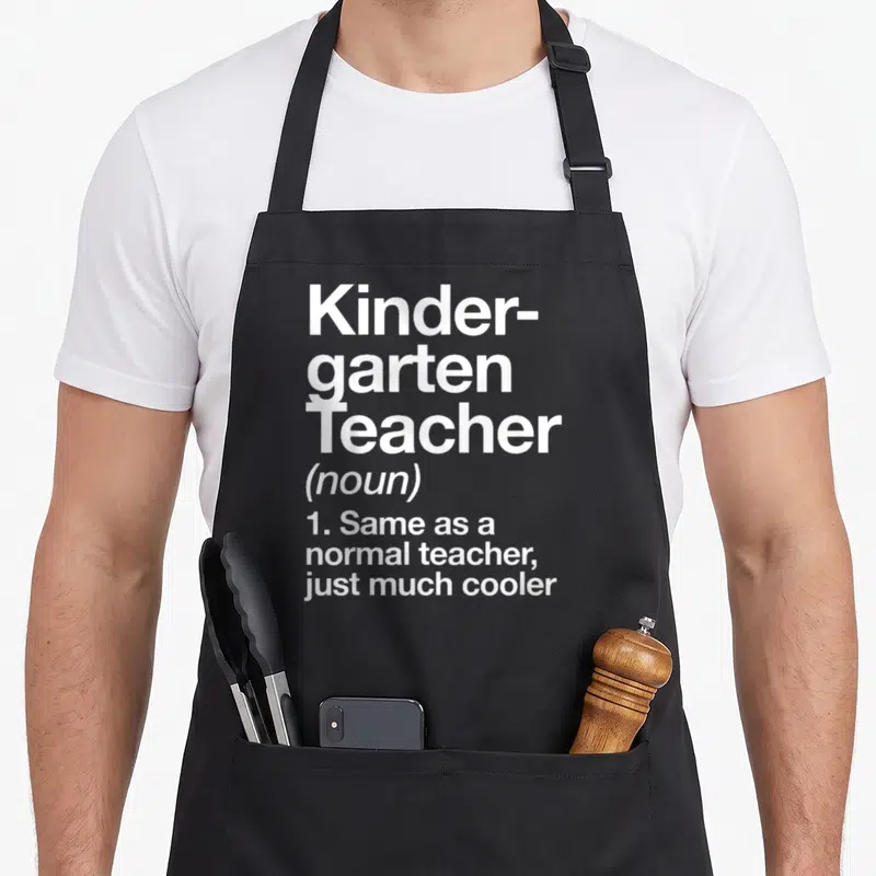 Definition kindergarten teacher apron - White stacked text reading Kinder-garten Teacher noun same as normal teacher — shown on a black cotton canvas apron against a white background