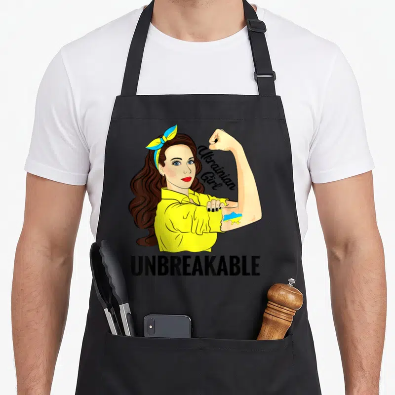 Unbreakable ukrainian aprons - Cartoon woman in yellow shirt flexing arm with Ukrainian map tattoo and UNBREAKABLE text — shown on a black cotton canvas apron against a white background