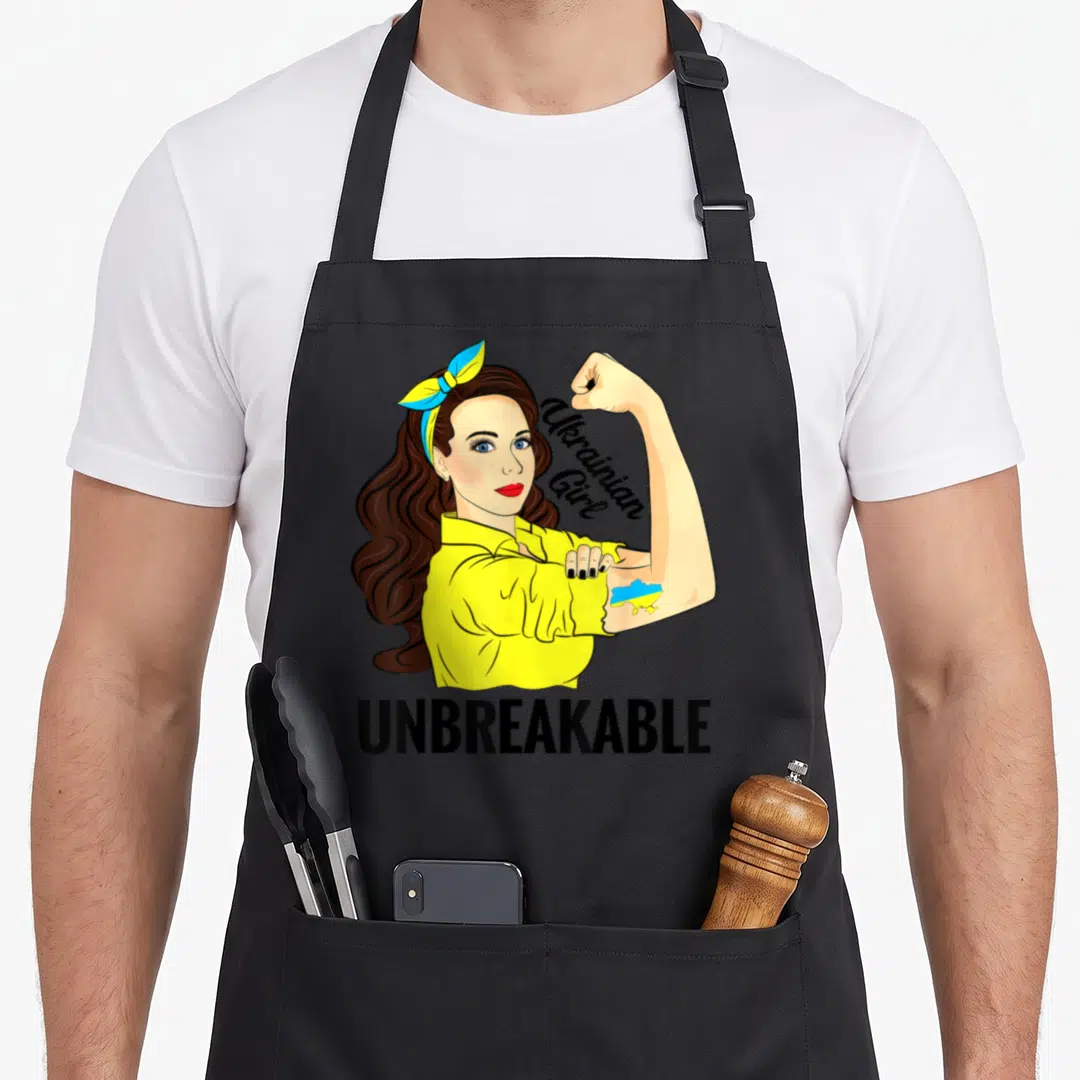 Unbreakable ukrainian aprons - Cartoon woman in yellow shirt flexing arm with Ukrainian map tattoo and UNBREAKABLE text — shown on a black cotton canvas apron against a white background