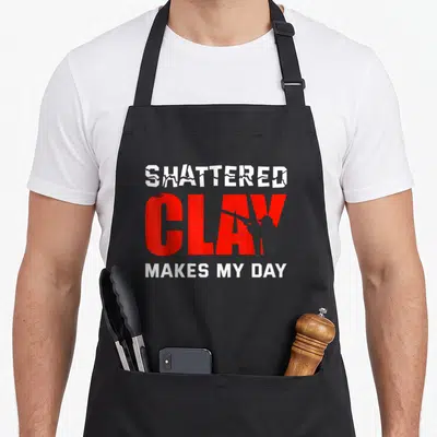 Shattered clay apron - Red and white text SHATTERED CLAY MAKES MY DAY with grey shooter silhouette inside the lettering — shown on a black cotton canvas apron against a white background