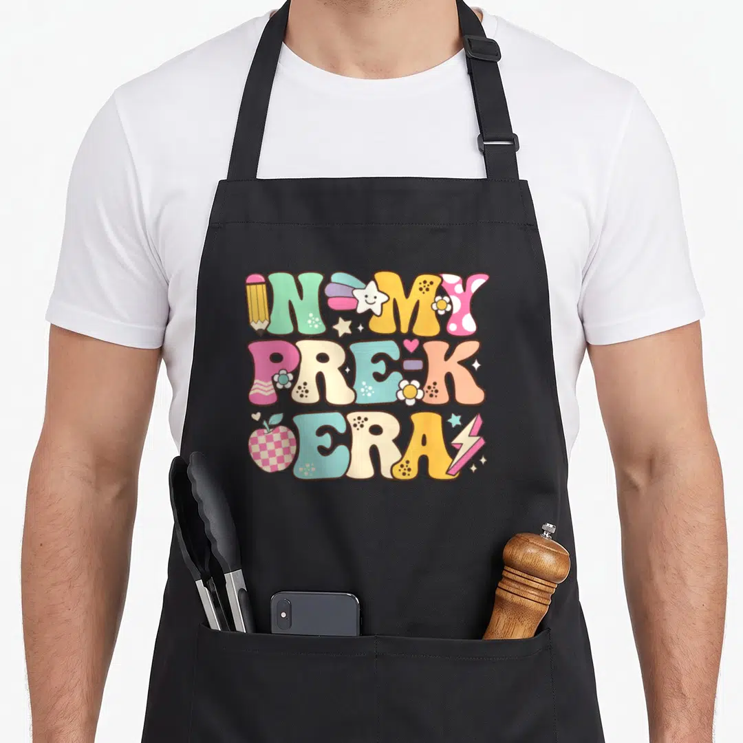 Era preschool teacher apron - Retro stacked text in my pre-k era with pencil, apple, star, and flower icons in pastel — shown on a black cotton canvas apron against a white background