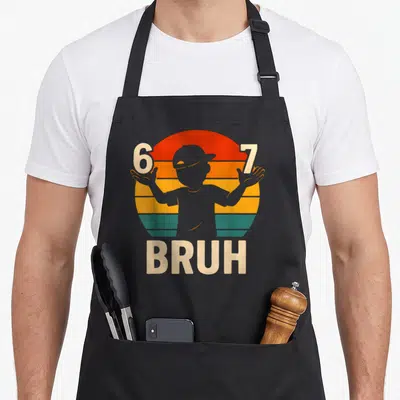 Kindergarten meme apron - Retro striped sun with 6 and 7 numbers, cap silhouette, and BRUH text below — shown on a black cotton canvas apron against a white background