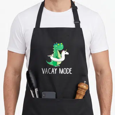 Mode dinosaur apron - Green dinosaur in white unicorn floatie with blue sunglasses and VACAY MODE text — shown on a black cotton canvas apron against a white background