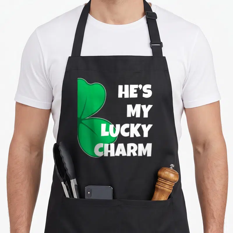 Patrick couple aprons - Green shamrock with white stacked text reading HE'S MY LUCKY CHARM — shown on a black cotton canvas apron against a white background