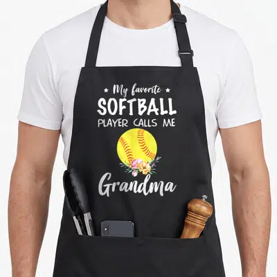 Softball player call grandma apron - Yellow softball with pink flowers and white text My favorite SOFTBALL PLAYER CALLS — shown on a black cotton canvas apron against a white background