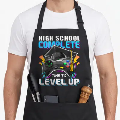 Complete video game apron - Game controller wearing a graduation cap with lightning bolts and completion text — shown on a black cotton canvas apron against a white background
