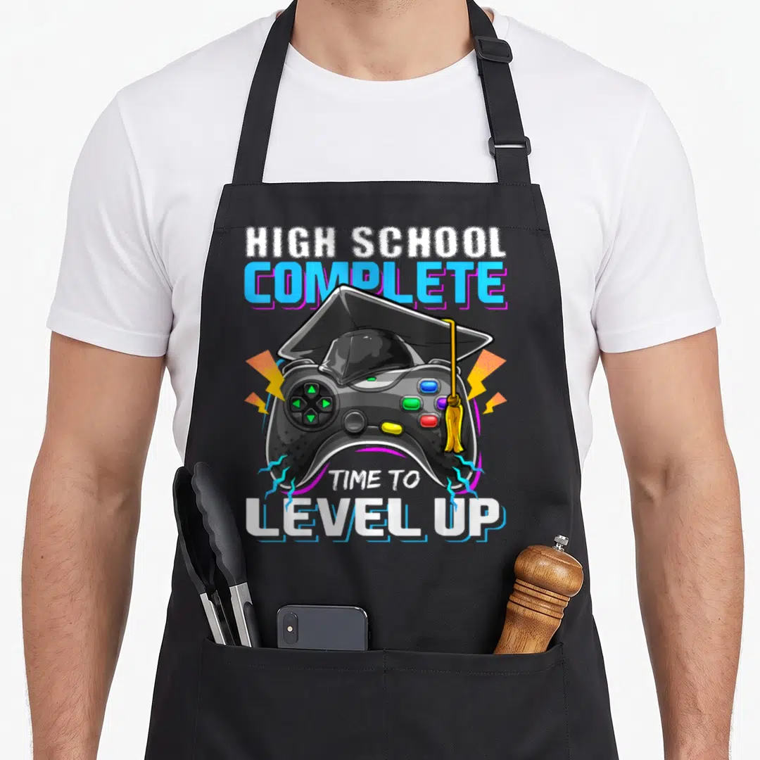 Complete video game apron - Game controller wearing a graduation cap with lightning bolts and completion text — shown on a black cotton canvas apron against a white background