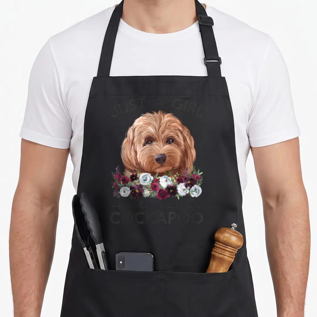 Cute cockapoo apron - Realistic golden brown cockapoo head with red and white flowers and arched text JUST A GIRL — shown on a black cotton canvas apron against a white background