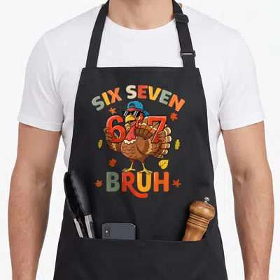 Meme thanksgiving apron - Cartoon turkey holding red six and seven digits with bruh text and autumn leaves — shown on a black cotton canvas apron against a white background