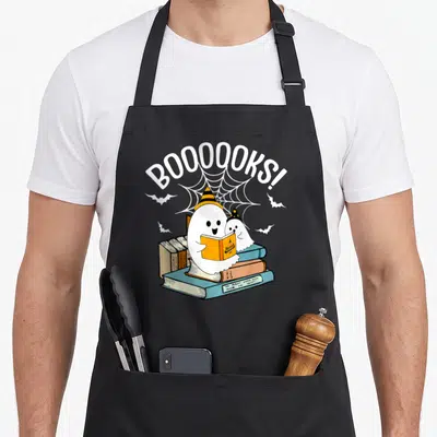 Booook ghost halloween apron - Two cartoon ghosts in witch hats reading books under arched BOOOOOKS! text and spider web — shown on a black cotton canvas apron against a white background