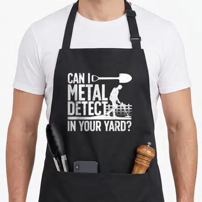 Vintage detectorist metal detecting apron - Distressed white text Can I Metal Detect In Your Yard with shovel — shown on a black cotton canvas apron against a white background