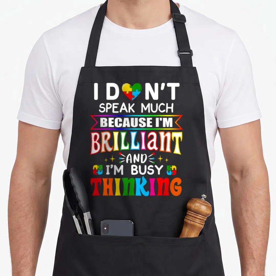 Speak autism awareness apron - Stacked white text I DON'T SPEAK MUCH BRILLIANT AND I'M BUSY THINKING with puzzle heart — shown on a black cotton canvas apron against a white background