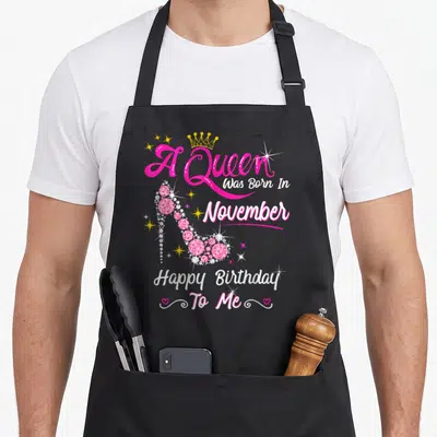 Cute queen november birthday apron - Pink high heel shoe with A Queen Was Born In November text and yellow crown — shown on a black cotton canvas apron against a white background