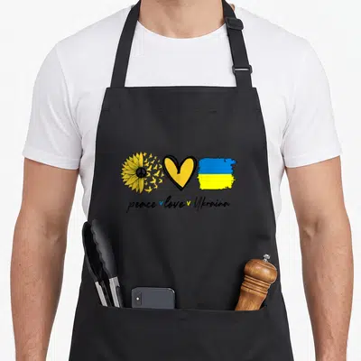 Peace sunflower apron - Sunflower with birds, heart, and flag above peace love Ukraina text — shown on a black cotton canvas apron against a white background