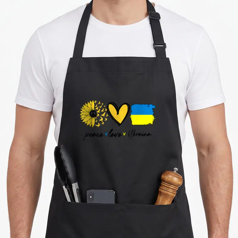 Peace sunflower apron - Sunflower with birds, heart, and flag above peace love Ukraina text — shown on a black cotton canvas apron against a white background