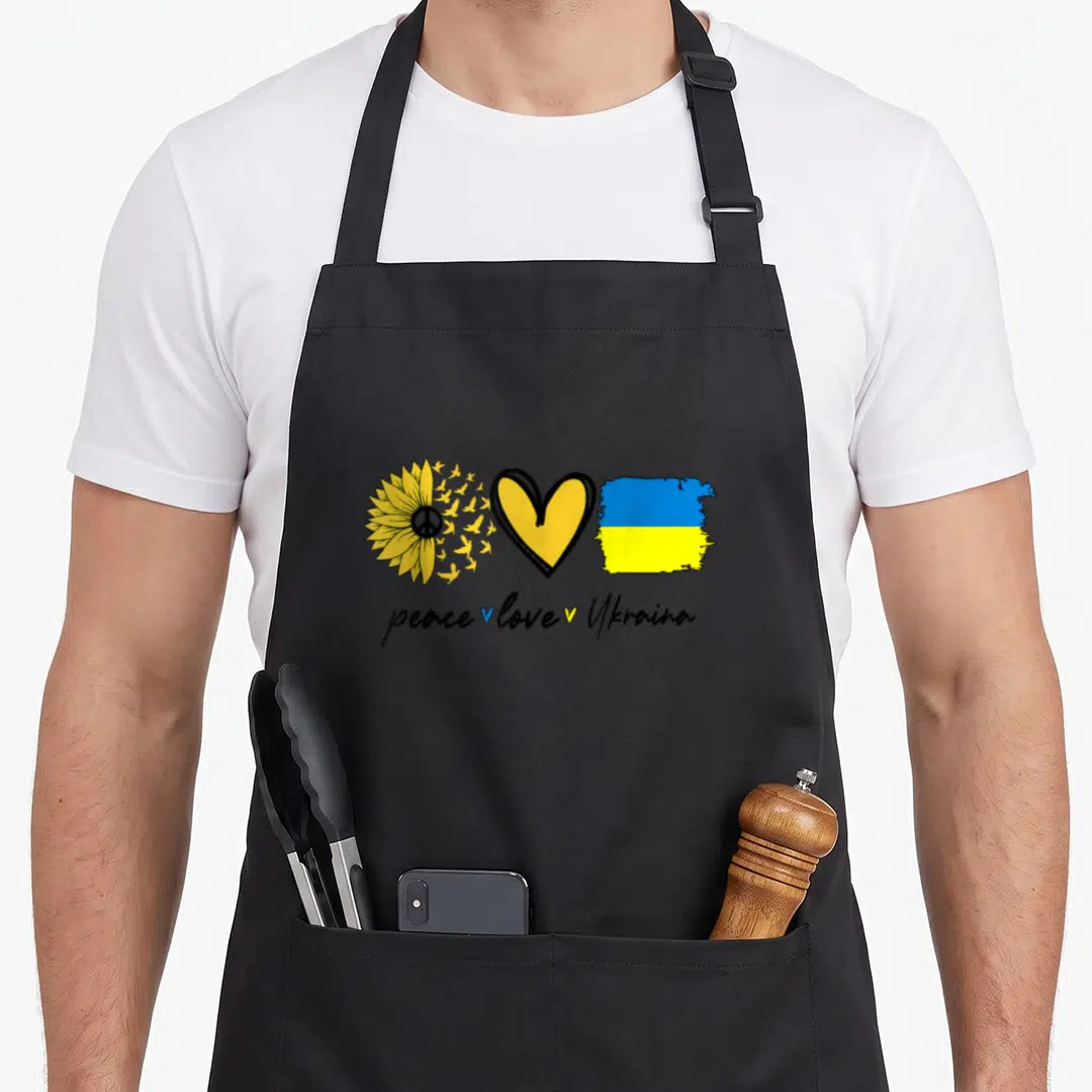 Peace sunflower apron - Sunflower with birds, heart, and flag above peace love Ukraina text — shown on a black cotton canvas apron against a white background
