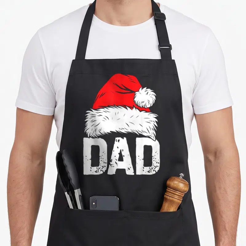 Family santa apron - Red and white Santa hat above distressed white DAD text on black background — shown on a black cotton canvas apron against a white background