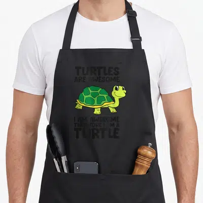 Funny turtle therefore apron - Cartoon green turtle flanked by black text reading Turtles Are Awesome I Am Awesome — shown on a black cotton canvas apron against a white background