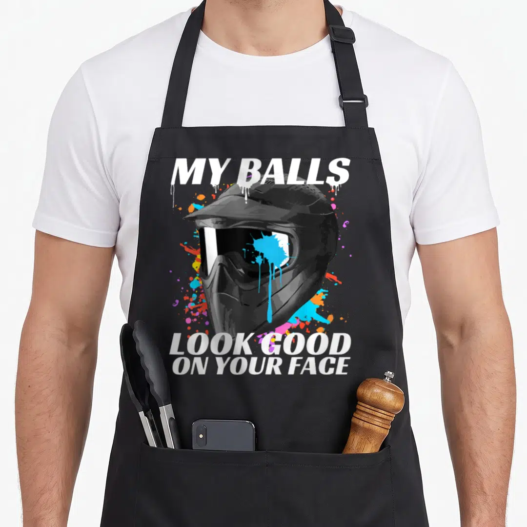 Paintballing face apron - Realistic black helmet with blue orange pink splatters and white dripping text — shown on a black cotton canvas apron against a white background