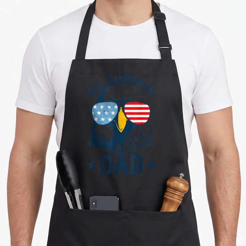 Daddy 4th of july apron - Bald eagle in flag sunglasses with ALL AMERICAN DAD text and two stars — shown on a black cotton canvas apron against a white background
