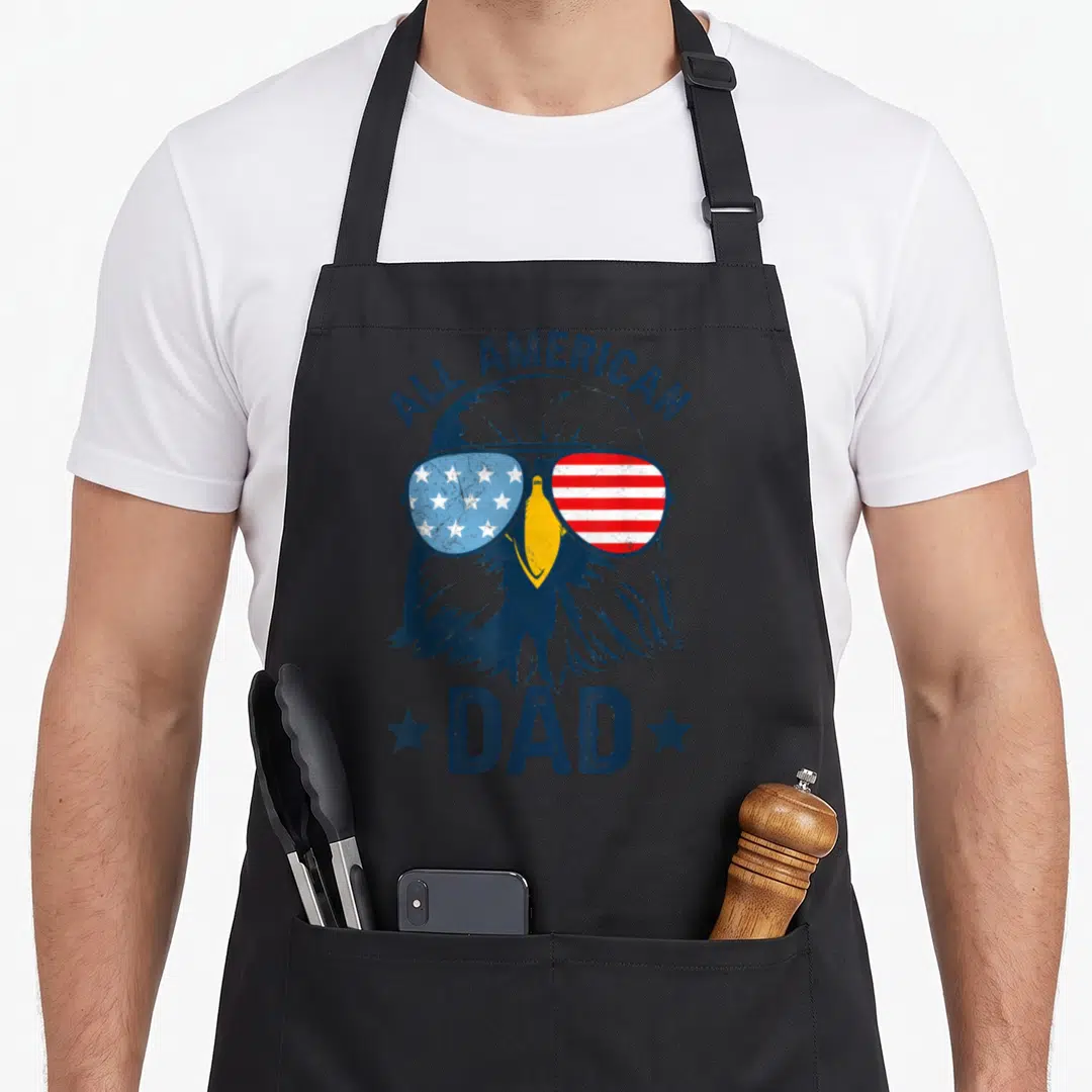 Daddy 4th of july apron - Bald eagle in flag sunglasses with ALL AMERICAN DAD text and two stars — shown on a black cotton canvas apron against a white background
