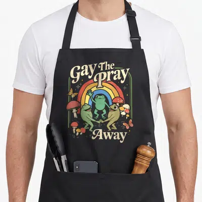 Gay frog aprons - Three green frogs dancing under a rainbow with Gay The Pray Away text and mushrooms — shown on a black cotton canvas apron against a white background