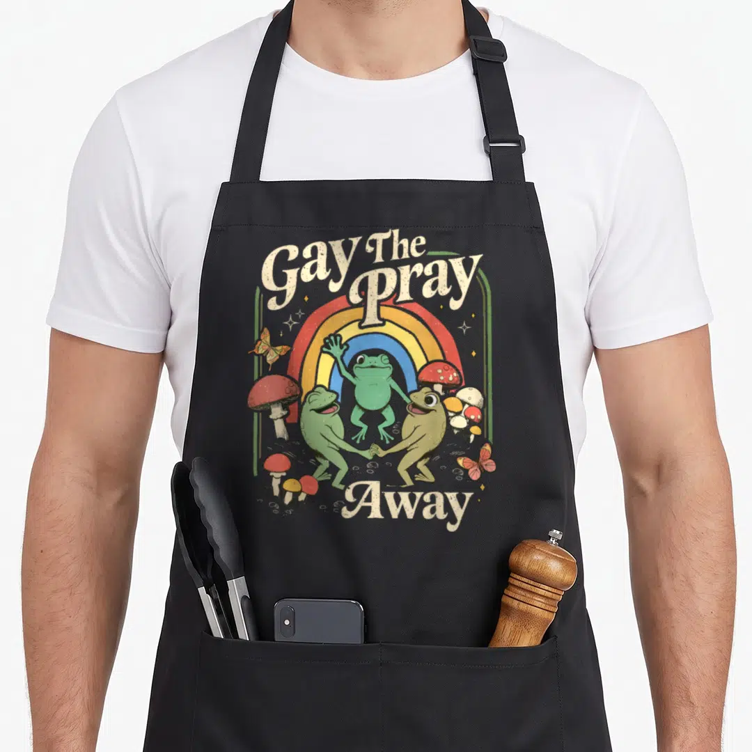 Gay frog aprons - Three green frogs dancing under a rainbow with Gay The Pray Away text and mushrooms — shown on a black cotton canvas apron against a white background